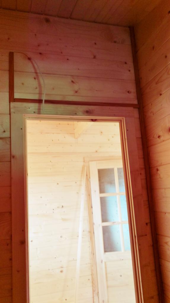 Wnętrze drewnianego domu w trakcie budowy z widocznym otworem na drzwi i wystającym kablem elektrycznym w górnej części ściany.