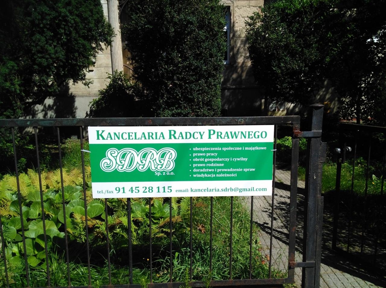 Tablica informacyjna Kancelarii Radcy Prawnego SDRB Sp. z o.o. umieszczona na metalowym ogrodzeniu, z wykazem oferowanych usług prawnych i danymi kontaktowymi, otoczona zielenią.