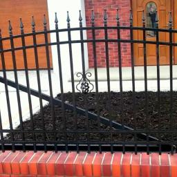 Micron-Tech - Czarny, metalowy płot z dekoracyjnymi elementami, osadzony na niskim murku z czerwonej cegły, na tle domu z cegły i drewnianej bramy garażowej.