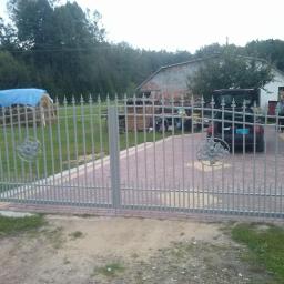 Micron-Tech - Szara, metalowa brama z ozdobnymi rozetami na tle podjazdu z kostki, w tle samochód z otwartym bagażnikiem i osoba przy budynku.