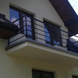 Micron-Tech - Balkon z czarną, metalową balustradą o prostym wzorze, widok z dołu na fragment elewacji domu z beżowym tynkiem.