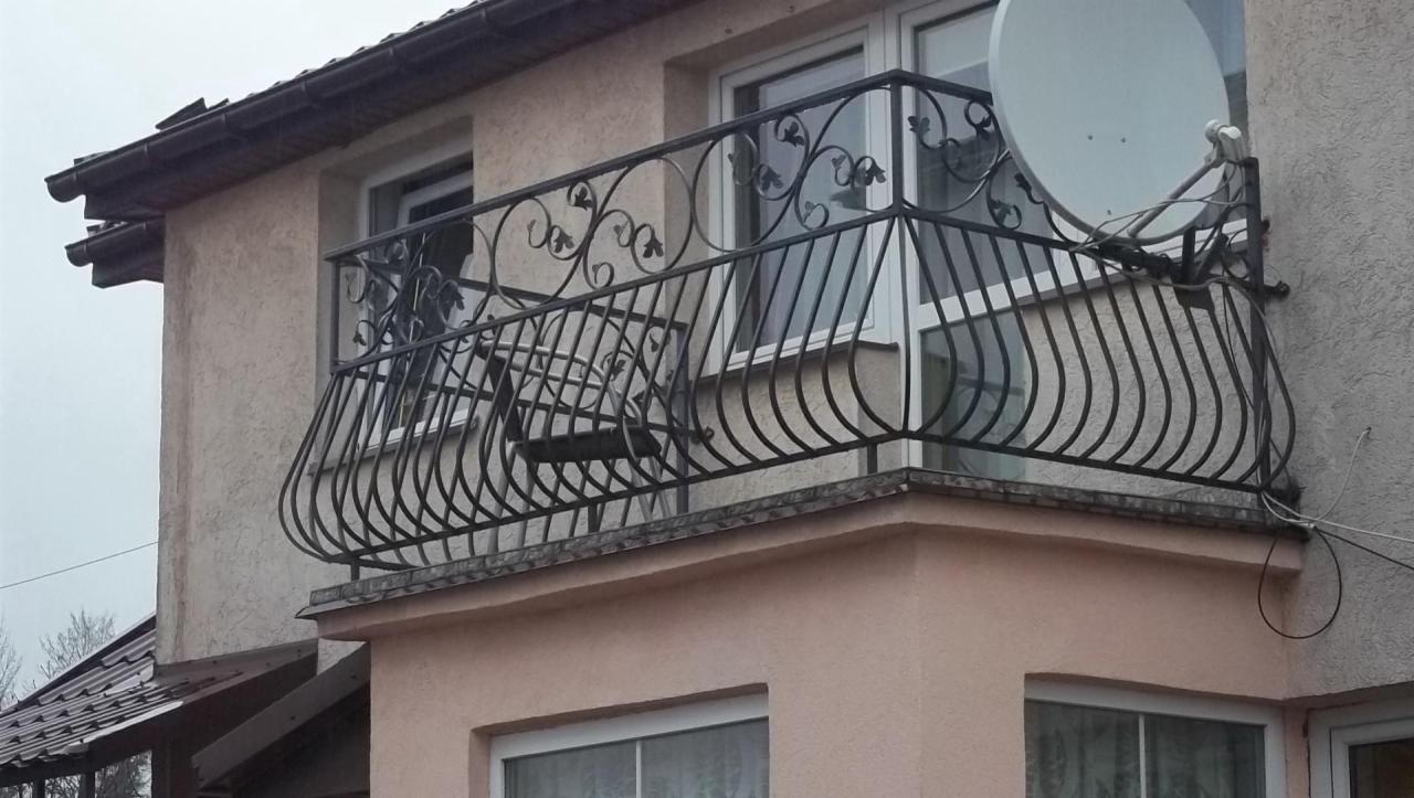 Kuty balkon z dekoracyjnymi elementami roślinnymi i anteną satelitarną, zamontowany na elewacji budynku z widocznymi oknami.