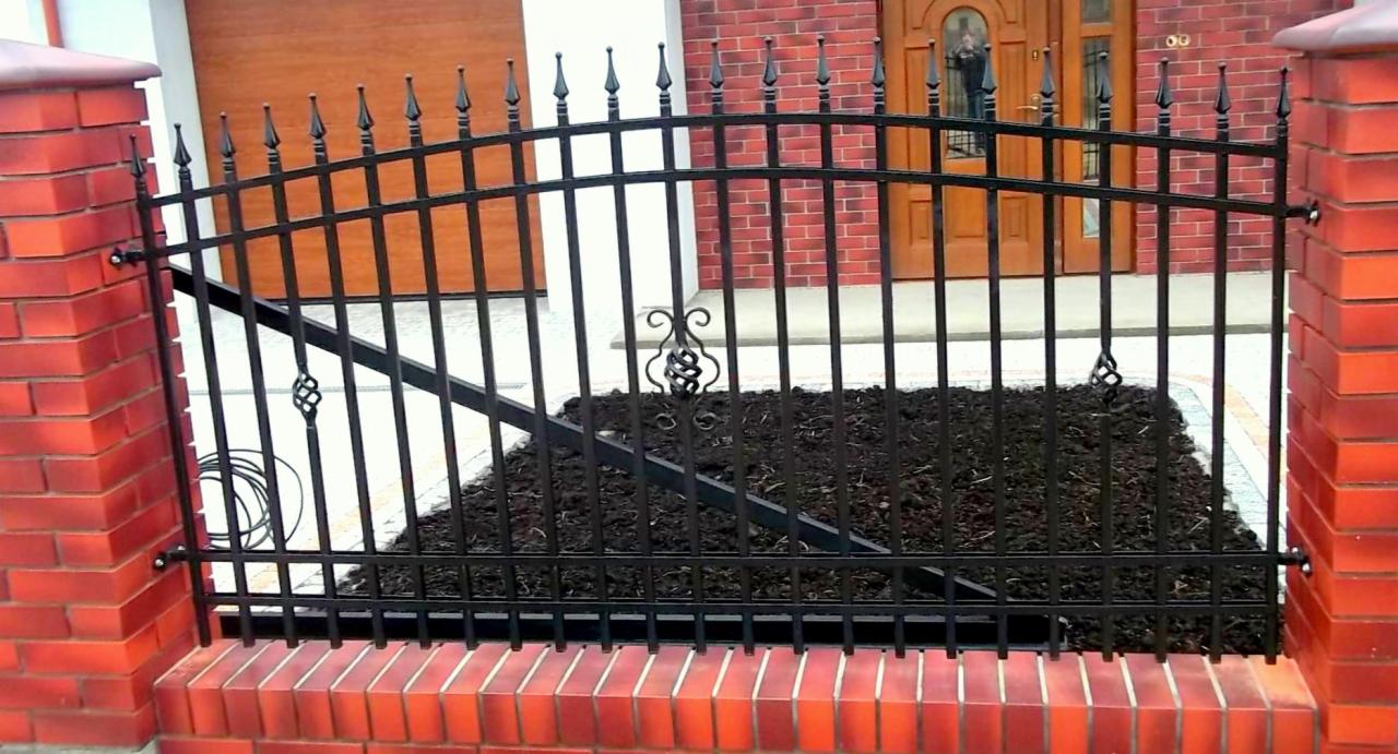 Czarny, metalowy płot z dekoracyjnymi elementami, osadzony na niskim murku z czerwonej cegły, na tle domu z cegły i drewnianej bramy garażowej.