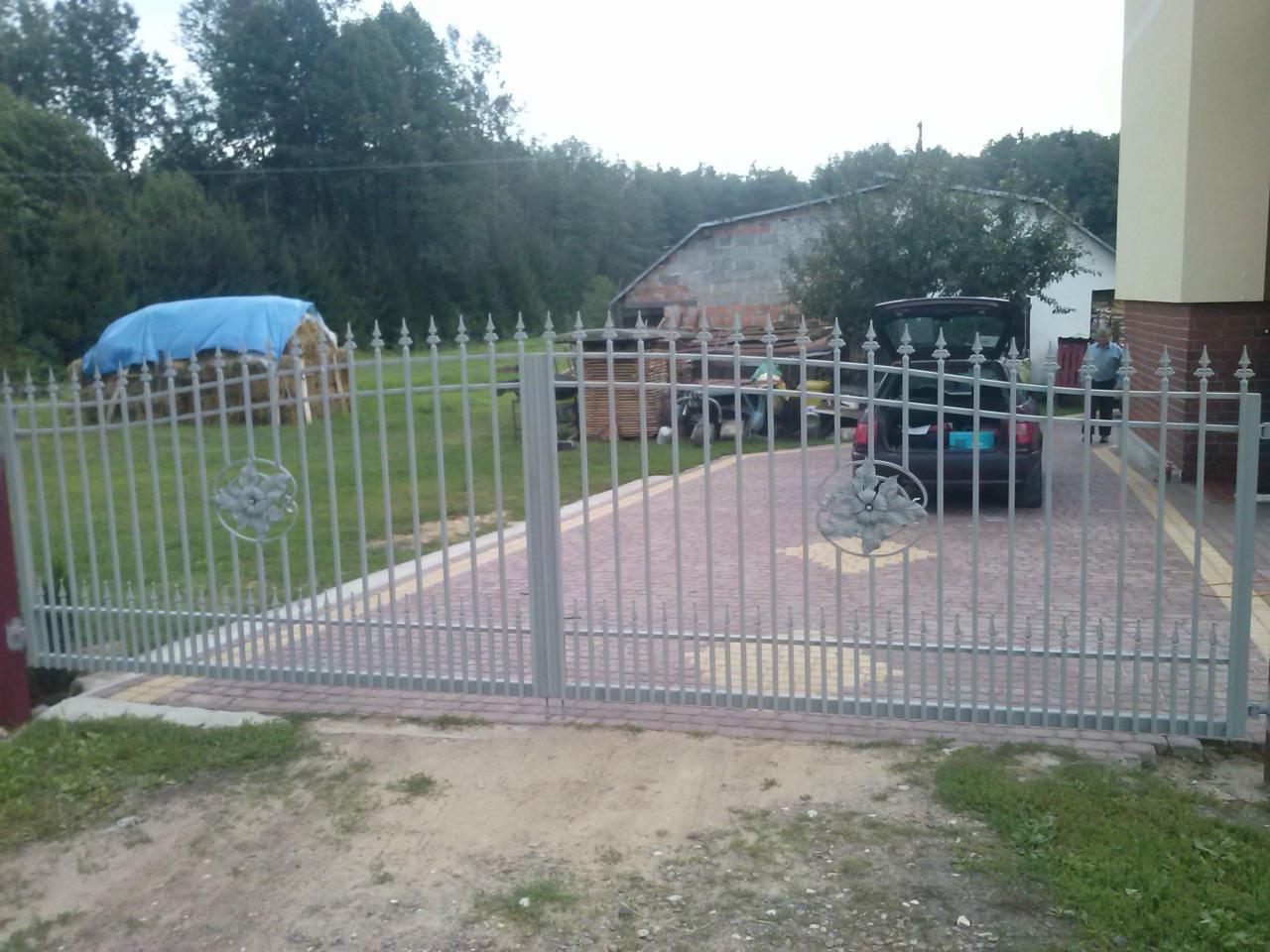 Szara, metalowa brama z ozdobnymi rozetami na tle podjazdu z kostki, w tle samochód z otwartym bagażnikiem i osoba przy budynku.