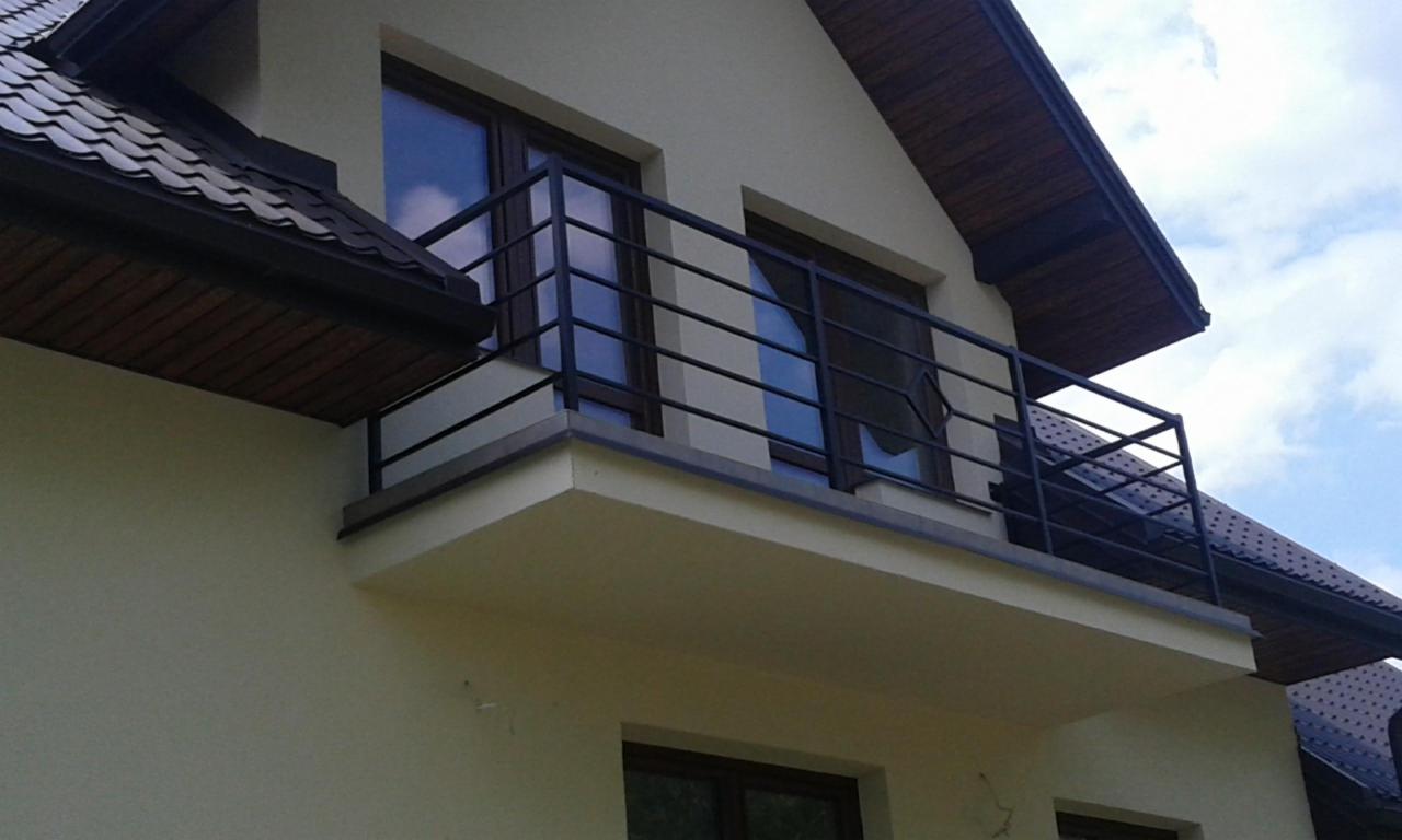Balkon z czarną, metalową balustradą o prostym wzorze, widok z dołu na fragment elewacji domu z beżowym tynkiem.