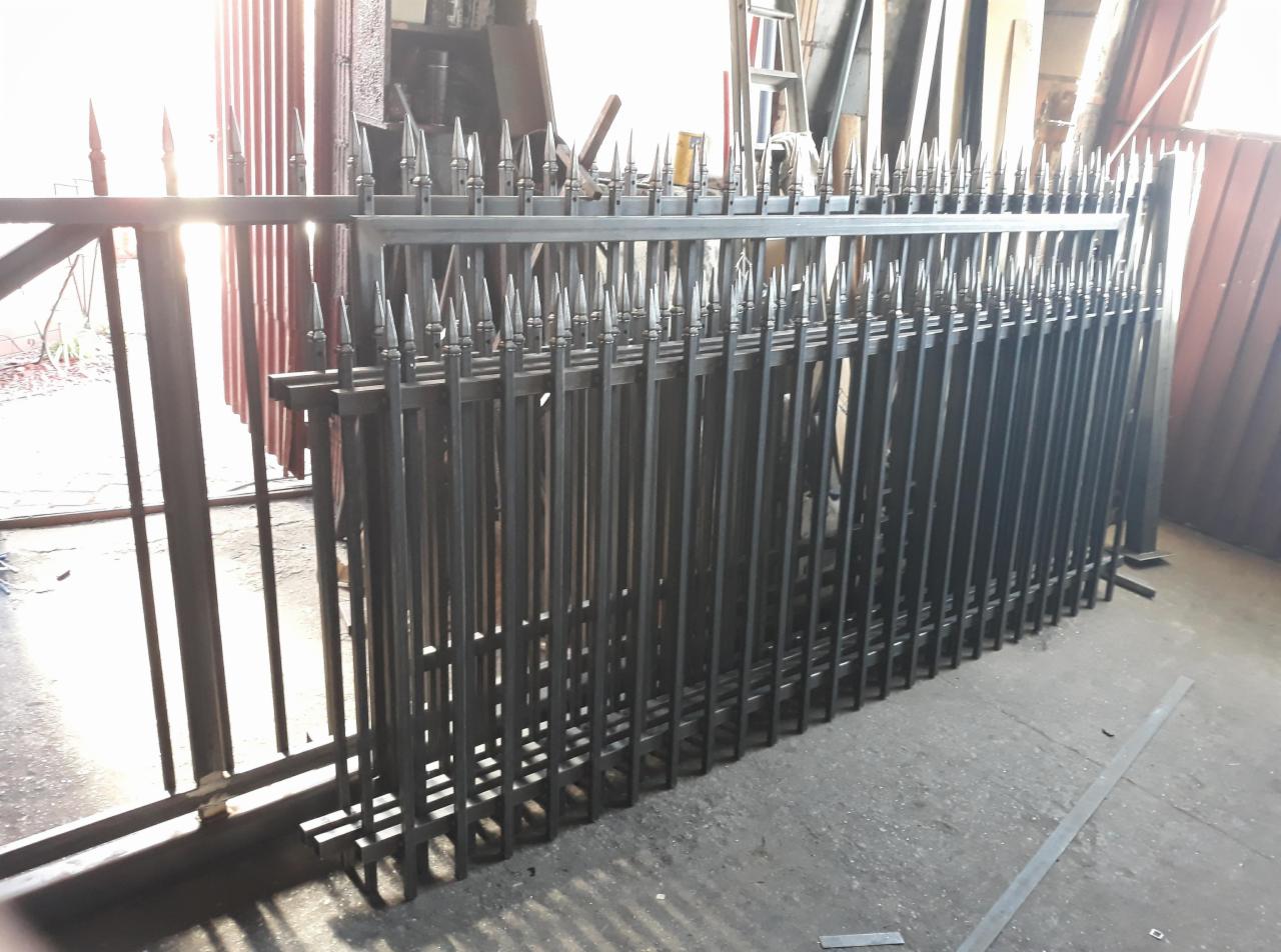 Stos czarnych, metalowych przęseł ogrodzeniowych z grotami, ułożonych jeden za drugim w warsztacie, widoczne narzędzia i elementy konstrukcyjne w tle.