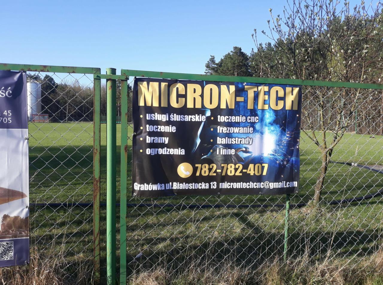Baner reklamowy firmy Micron-Tech z ofertą usług ślusarskich, toczonych CNC, bram, ogrodzeń i balustrad, zawieszony na ogrodzeniu z siatki.