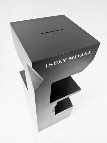 Metalowa konstrukcja spawana w Kamiennej Górze, przypominająca literę E z dodatkową skrzynką na górze z napisem 'ISSEY MIYAKE', w czarno-białej tonacji.