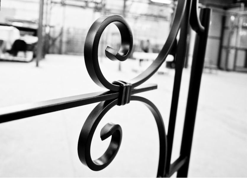 Czarno-białe zbliżenie fragmentu metalowej, ozdobnej balustrady z charakterystycznymi spiralnymi zdobieniami, widoczne elementy łączenia prętów, w tle hala produkcyjna.