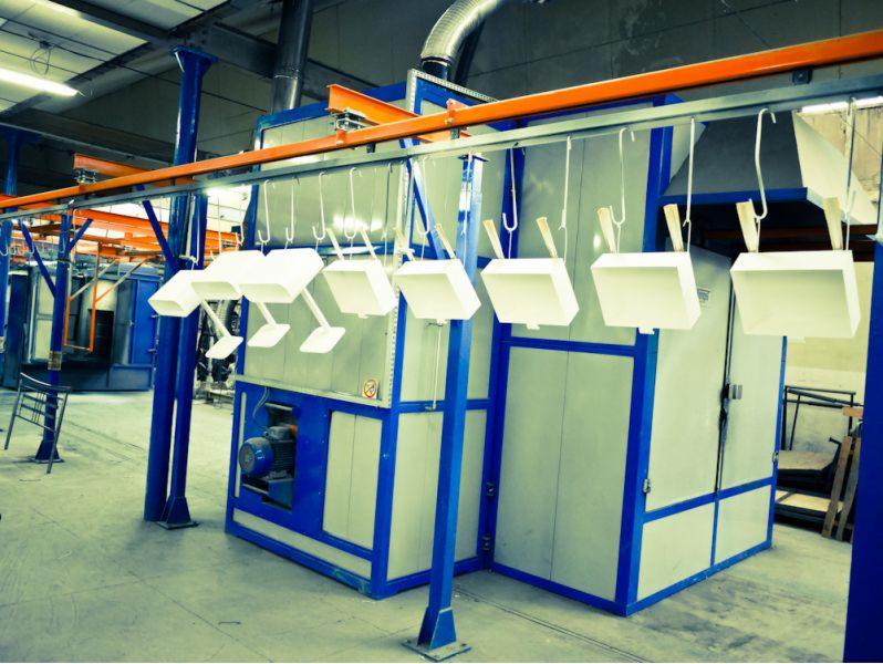 Elementy metalowe w trakcie malowania proszkowego, zawieszone na hakach przed piecem.