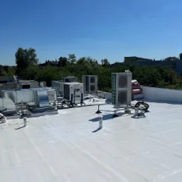 Biały, płaski dach pokryty hydroizolacją z widocznymi systemami wentylacji i klimatyzacji, na tle zieleni drzew i budynków, w słoneczny dzień.