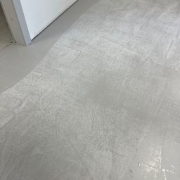 Purmaster - Świeżo wylana, szara posadzka przemysłowa z widocznymi śladami po narzędziach i smugami, w rogu pomieszczenia przy białej ścianie i futrynie drzwi.