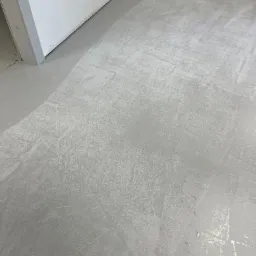 Świeżo wylana, szara posadzka przemysłowa z widocznymi śladami po narzędziach i smugami, w rogu pomieszczenia przy białej ścianie i futrynie drzwi.