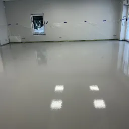 Błyszcząca, szara posadzka w nowym pomieszczeniu, zabezpieczone folią okna i ściany, widoczne odbicia światła w gładkiej powierzchni podłogi.