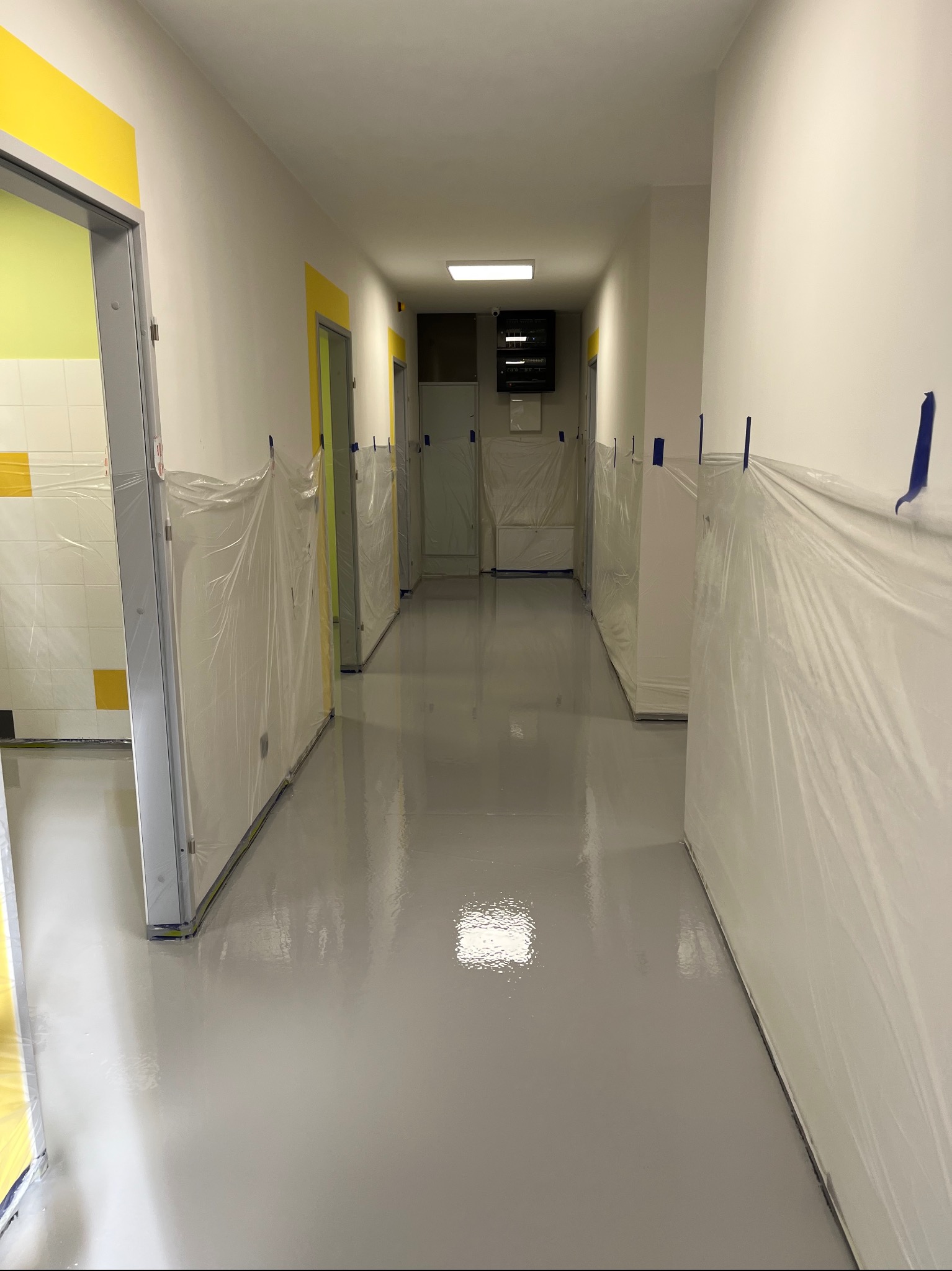 Korytarz w budynku użyteczności publicznej z nową, błyszczącą, szarą posadzką, zabezpieczone ściany i drzwi folią ochronną i taśmą malarską, widoczne elementy instalacji elektrycznej na ścianie.