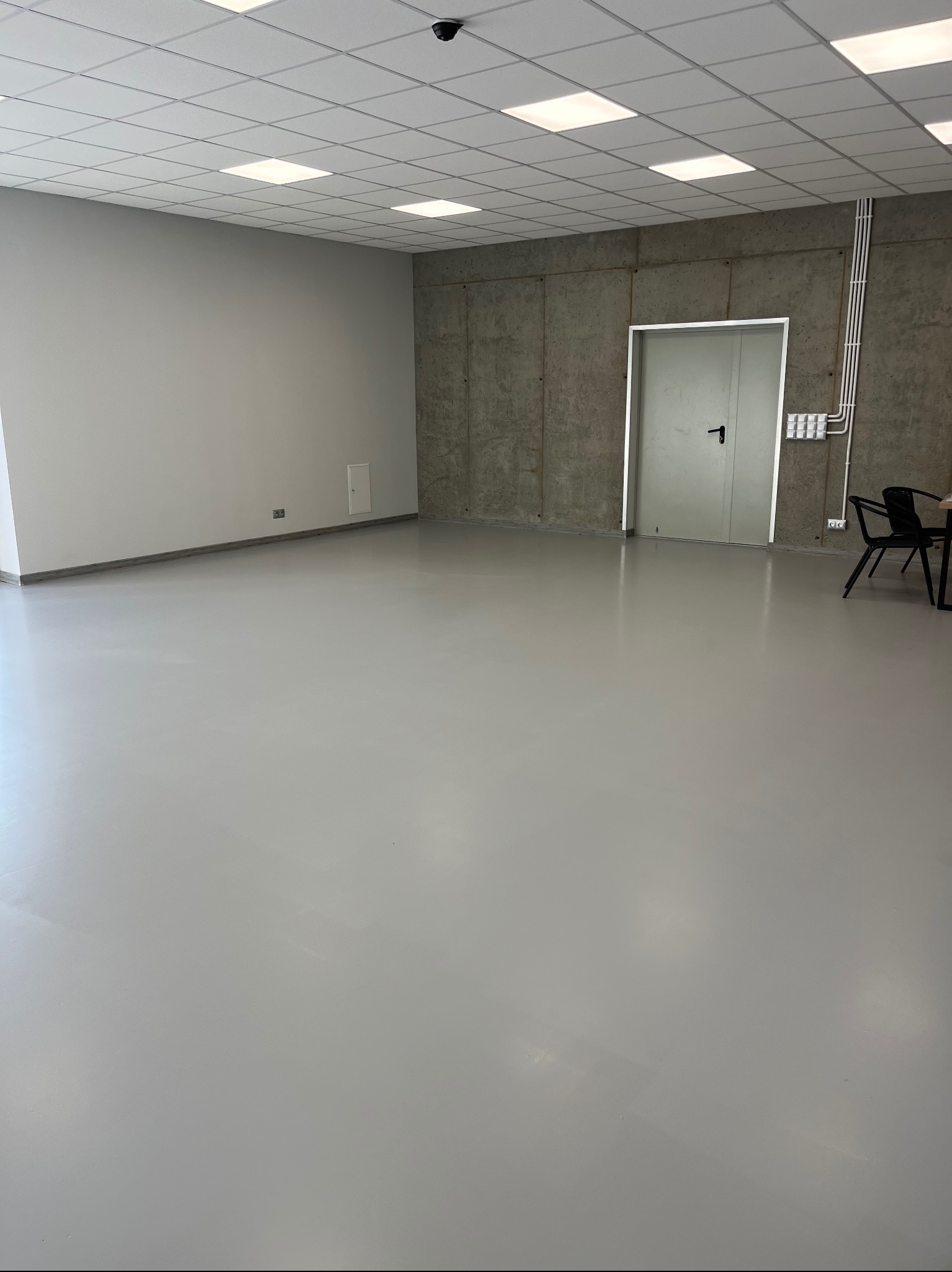 Jasnoszara, gładka posadzka przemysłowa w nowo wykończonym, przestronnym pomieszczeniu z betonowymi ścianami i podwieszanym sufitem kasetonowym.
