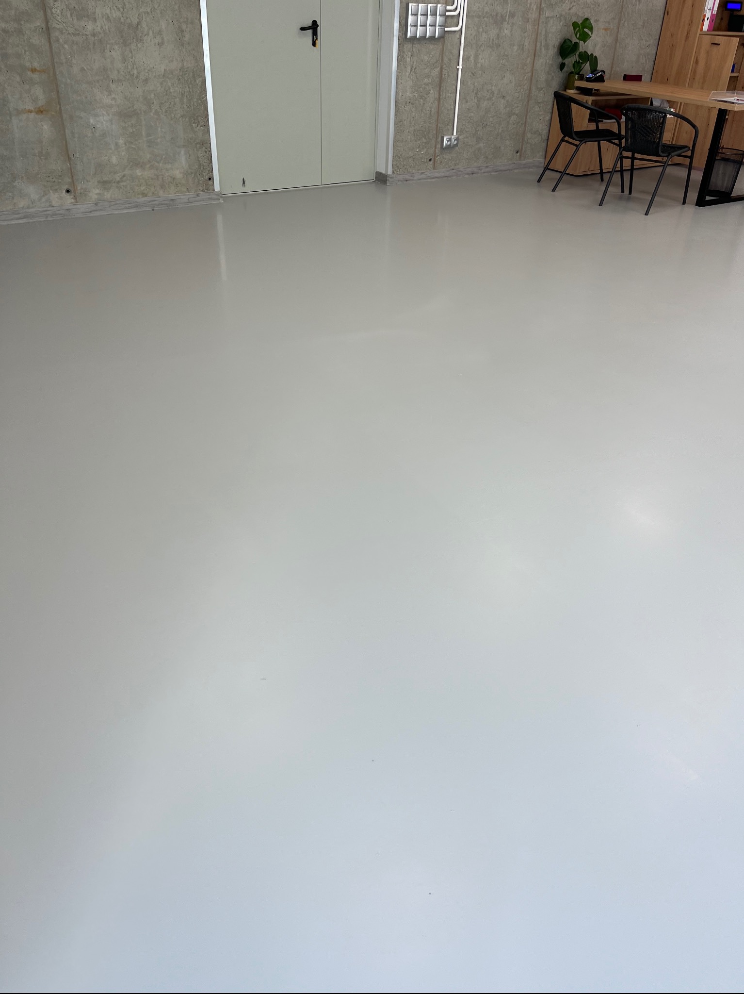 Jasnoszara, gładka posadzka w pomieszczeniu biurowym z betonowymi ścianami i minimalistycznym umeblowaniem, widoczne krzesła i biurko w tle.