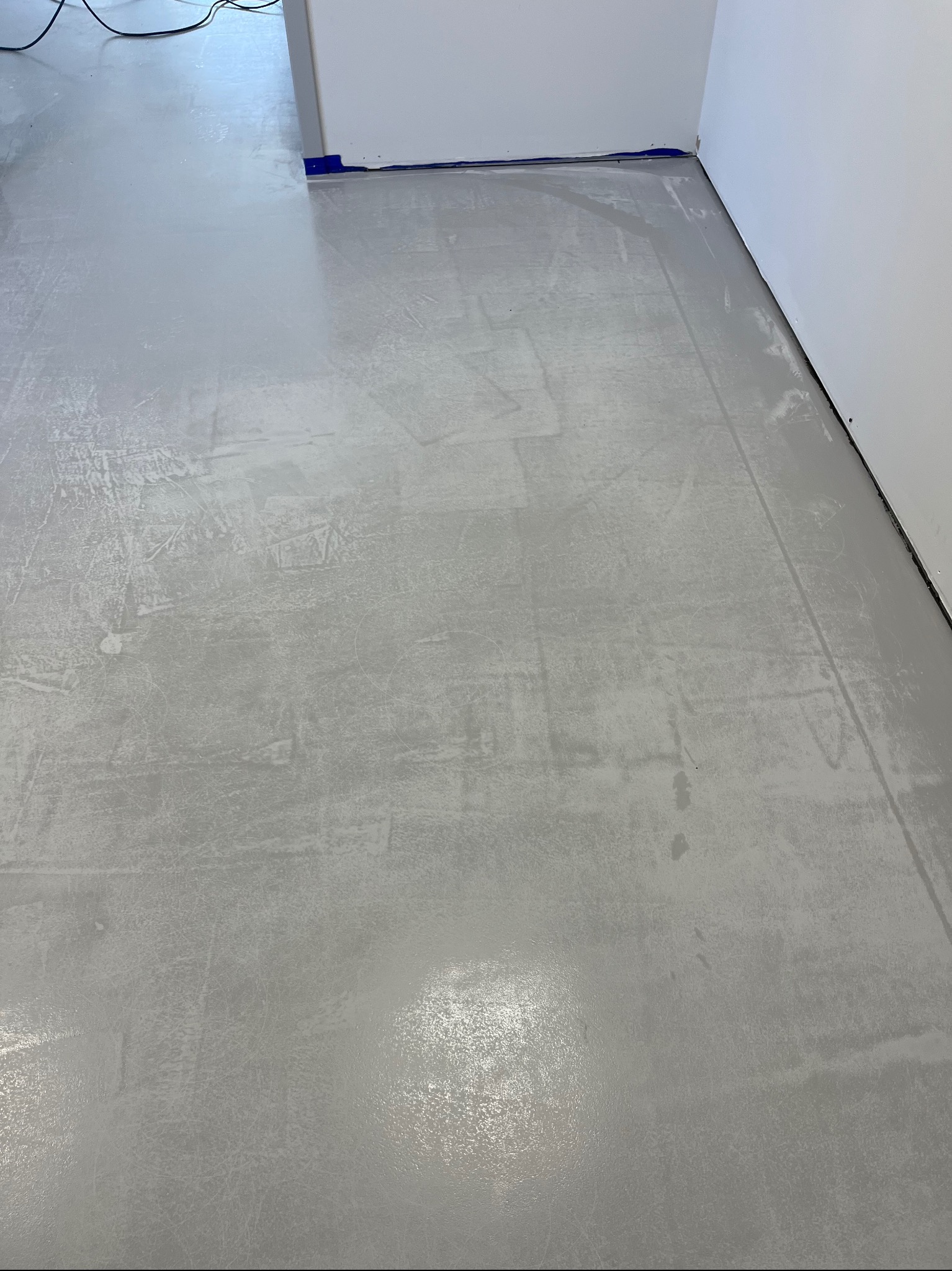 Świeżo wylana, jasnoszara posadzka z żywicy epoksydowej, jeszcze mokra i odbijająca światło, z widocznymi smugami i nierównościami powierzchni, zabezpieczona niebieską taśmą malarską przy ścianach.