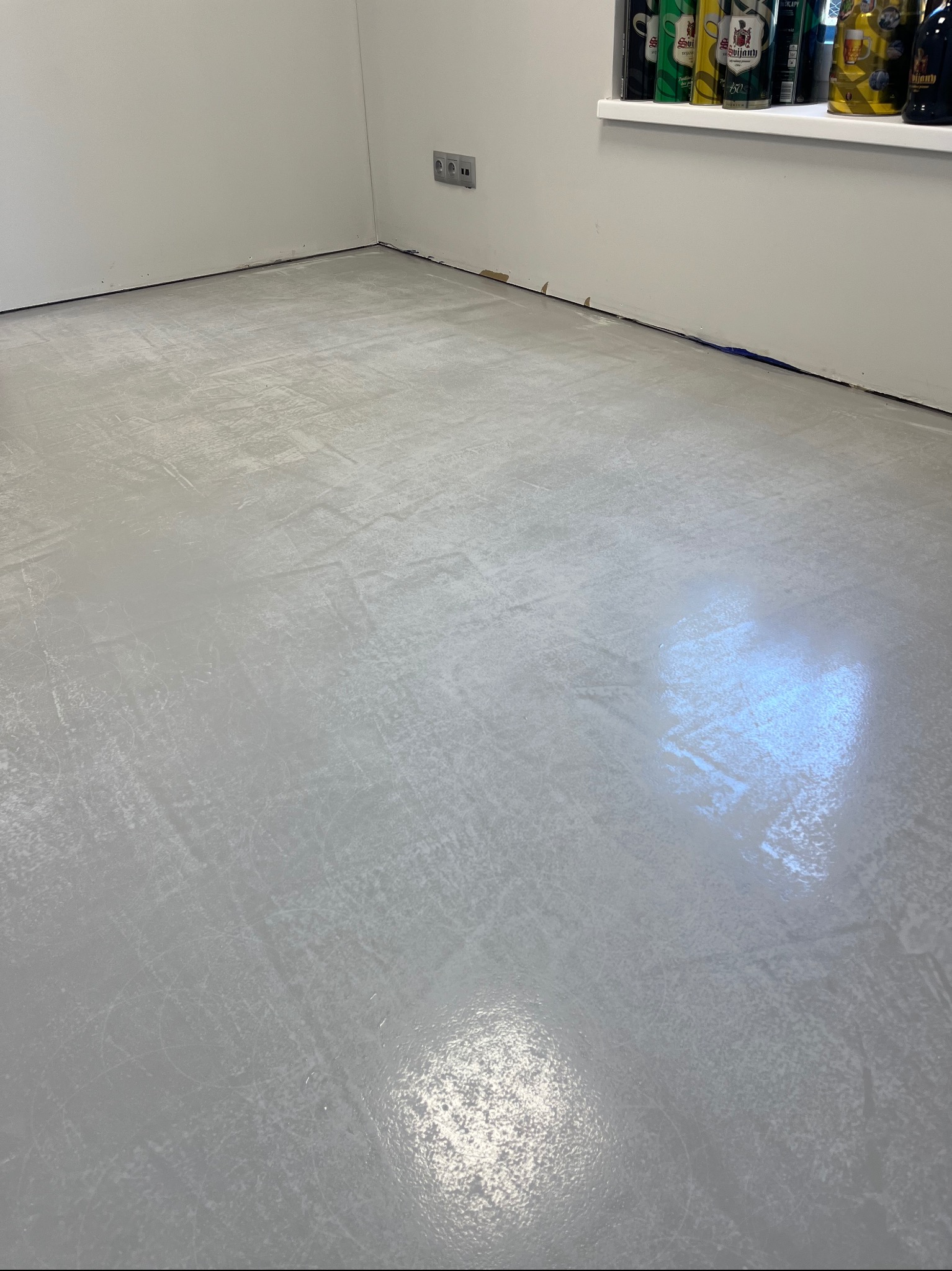 Świeżo wylana, jasnoszara posadzka żywiczna w pomieszczeniu z białymi ścianami, widoczne ślady po wałku malarskim i delikatne refleksy świetlne, przy ścianie rząd puszek na parapecie.