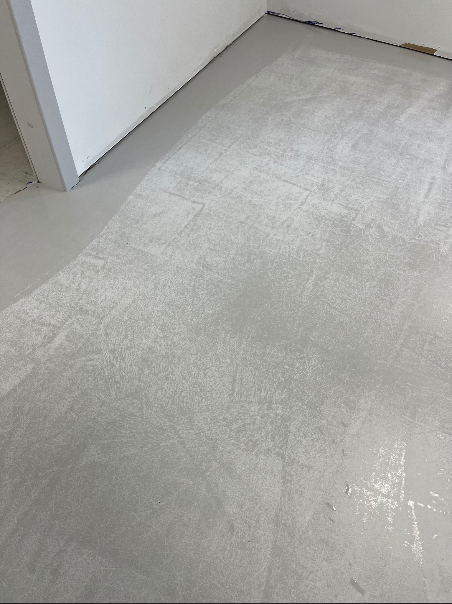 Świeżo wylana, szara posadzka przemysłowa z widocznymi śladami po narzędziach i smugami, w rogu pomieszczenia przy białej ścianie i futrynie drzwi.