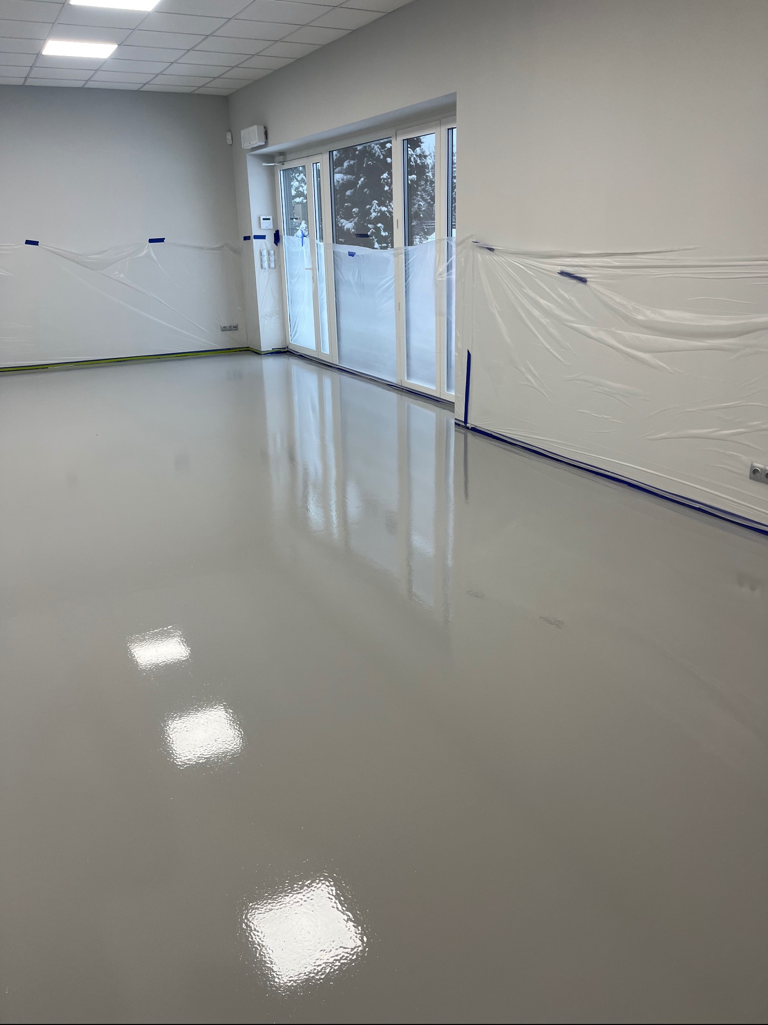 Świeżo wylana, gładka, szara posadzka żywiczna w jasnym pomieszczeniu, ściany zabezpieczone folią malarską, widoczne odbicia światła od lamp sufitowych.