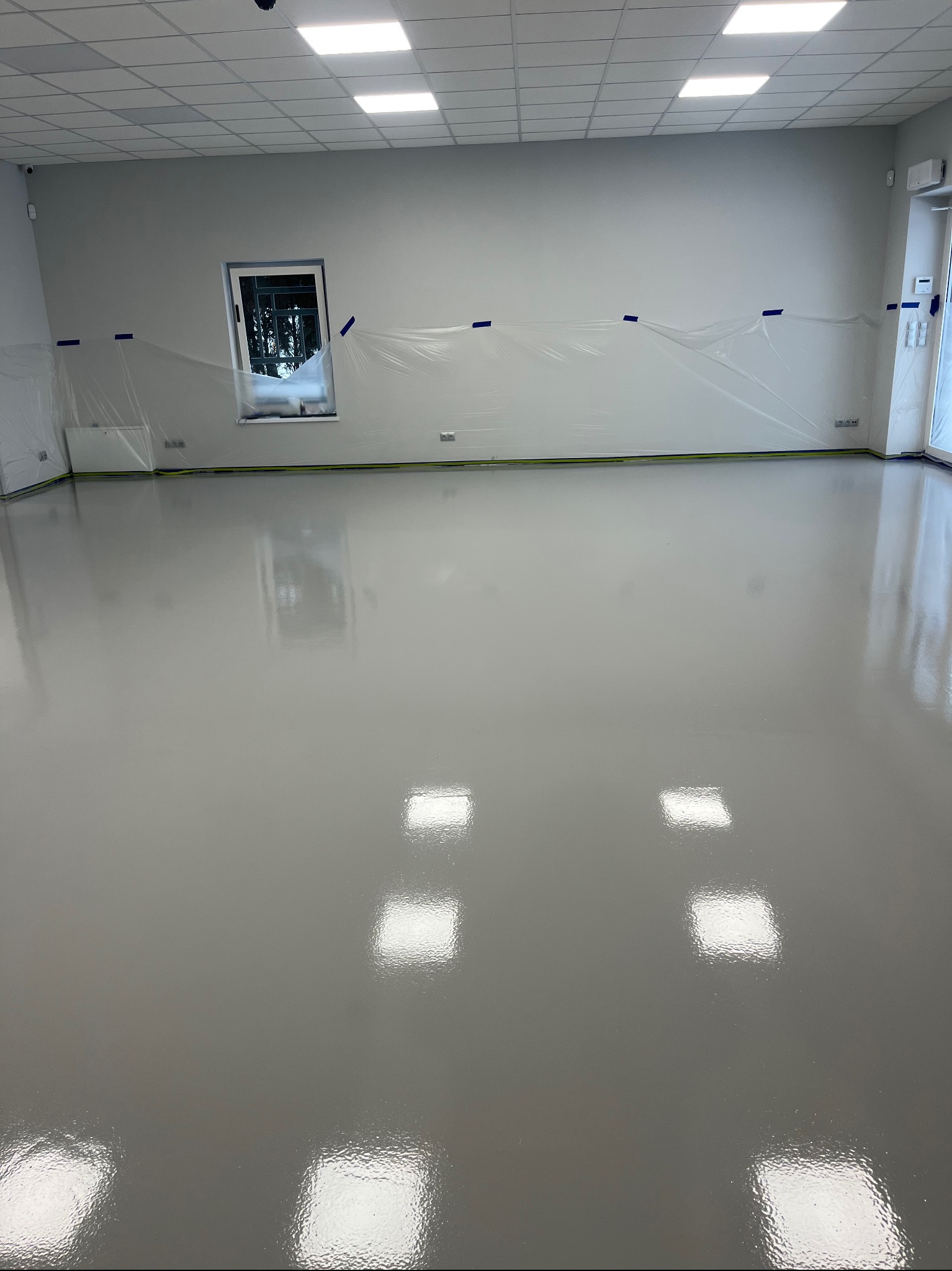 Błyszcząca, szara posadzka w nowym pomieszczeniu, zabezpieczone folią okna i ściany, widoczne odbicia światła w gładkiej powierzchni podłogi.