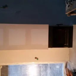 Wnętrze w trakcie wykańczania z obudowanym kominkiem, widoczne ślady szpachlowania i przygotowania pod malowanie, niezabezpieczone okno folią malarską oraz prowizoryczny stołek roboczy.