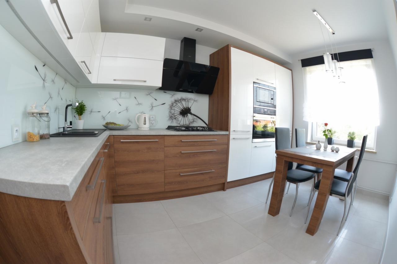 Jasna kuchnia z drewnianymi szafkami dolnymi i białymi górnymi, czarnym okapem i szklanym panelem z nadrukiem dmuchawców, uzupełniona drewnianym stołem z krzesłami.