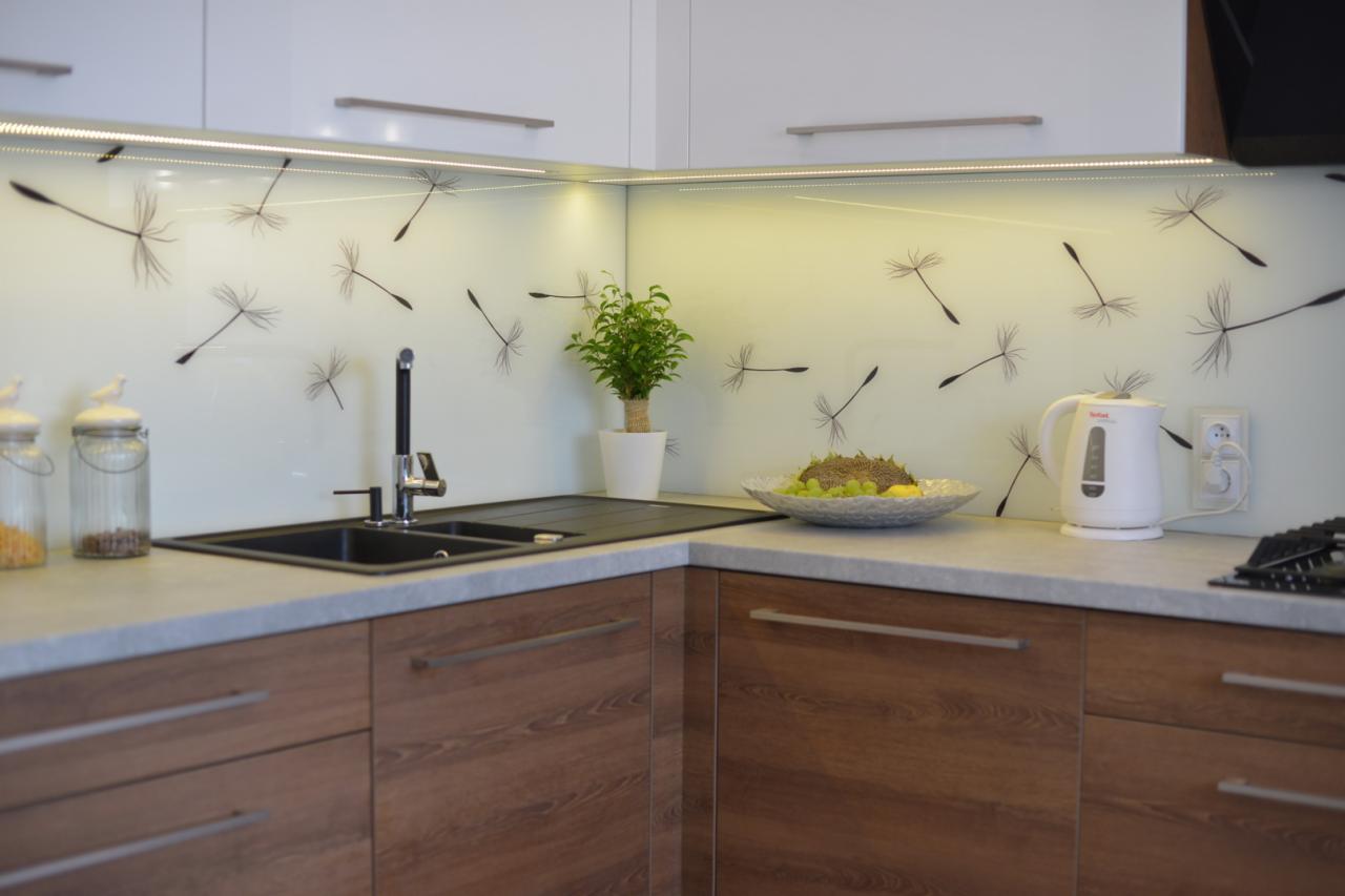 Nowoczesna kuchnia z drewnianymi frontami szafek, czarnym zlewozmywakiem i panelem szklanym z motywem dmuchawca, oświetlona listwą LED, z czajnikiem elektrycznym i misą owoców na blacie.