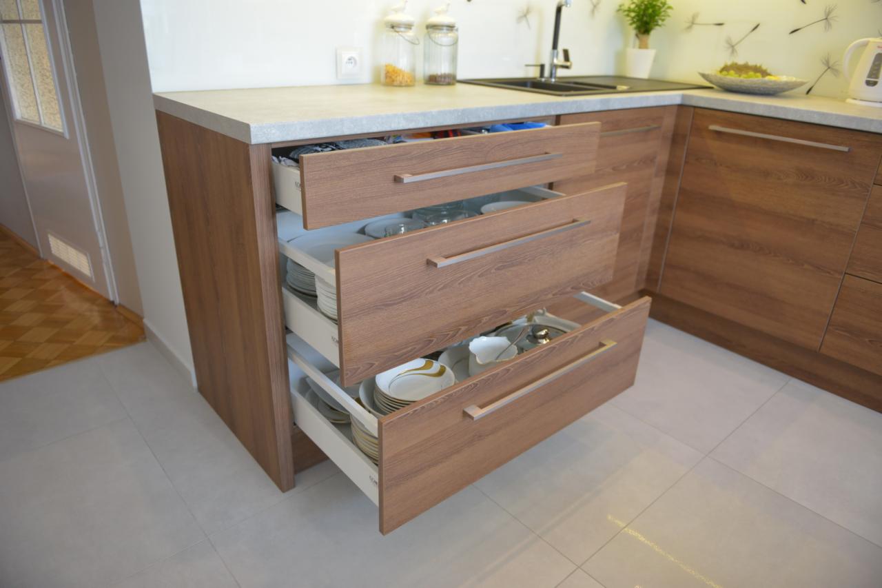 Kuchnia z drewnianymi frontami i wysuwanymi szufladami, w których widać naczynia, talerze i szklanki, z blatem w kolorze szarego betonu i białymi ścianami w tle.