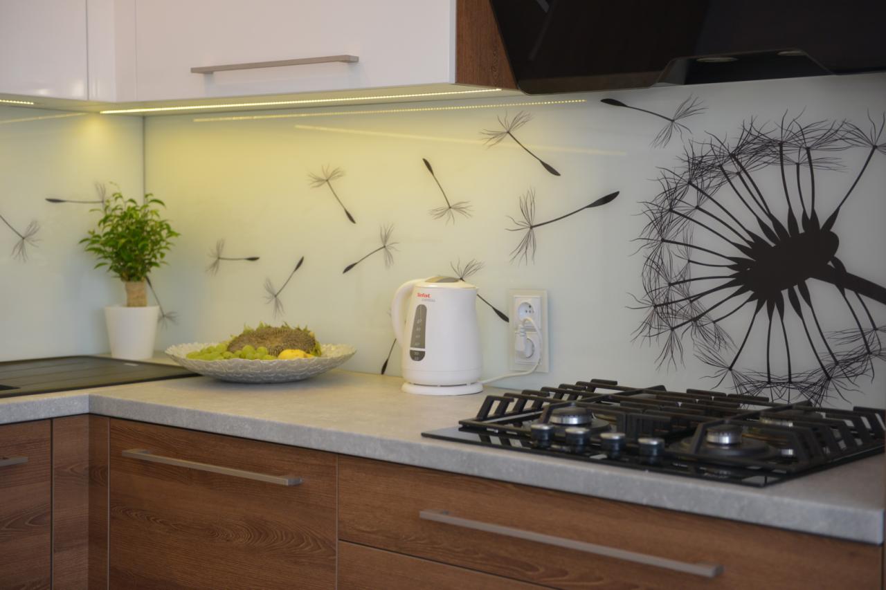 Nowoczesna kuchnia z drewnianymi frontami, blatem w odcieniach szarości i panelem szklanym z motywem dmuchawca, oświetlona listwą LED pod szafkami, z czajnikiem elektrycznym i płytą gazową.