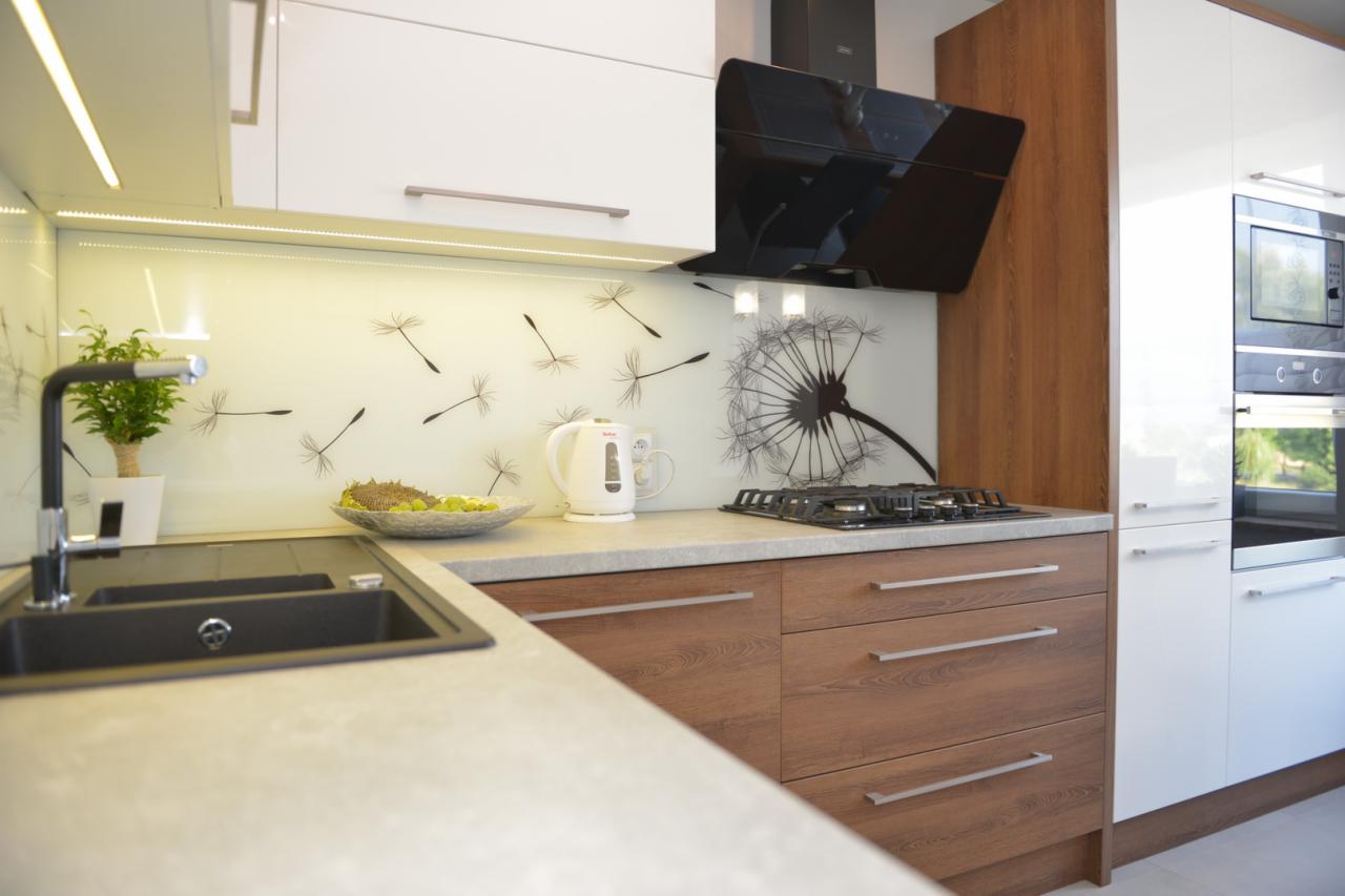 Nowoczesna kuchnia z białymi i drewnopodobnymi frontami, szklany panel nad blatem z motywem dmuchawca, czarny okap, zlewozmywak z baterią i dekoracyjna roślina w doniczce.