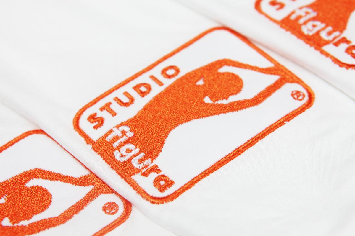 Białe tkaniny z pomarańczowym haftem logo 'Studio Figura', zbliżenie na detale ściegów i fakturę materiału.