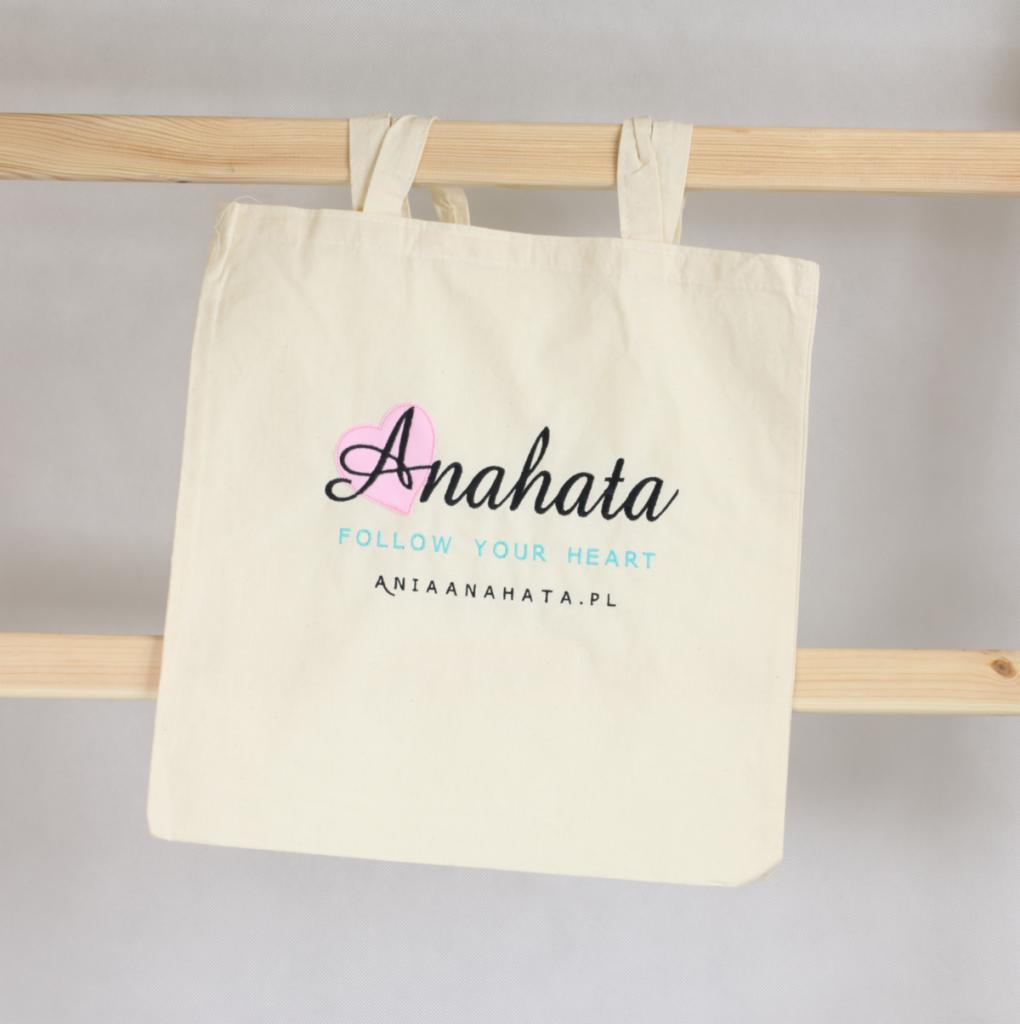 Beżowa torba bawełniana z czarnym napisem 'Anahata' z różowym sercem, hasłem 'Follow your heart' i adresem strony internetowej, zawieszona na drewnianej drabince.