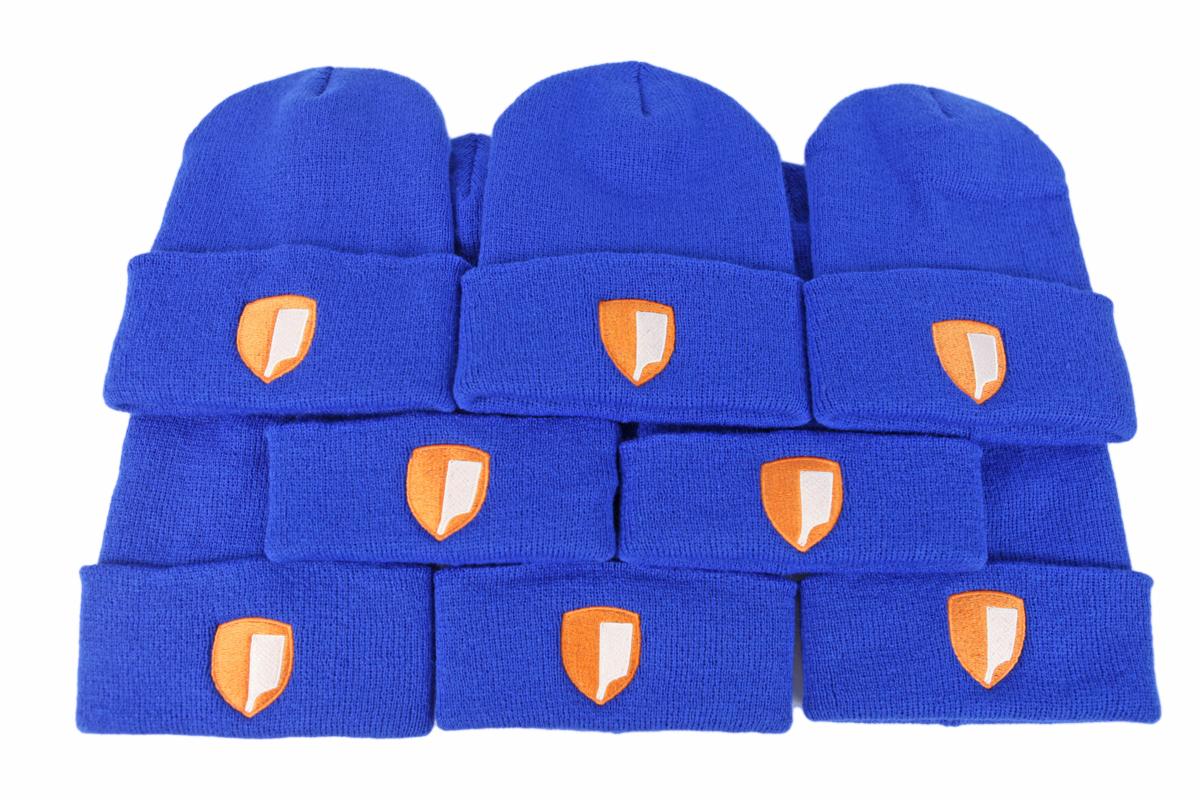 Stos niebieskich czapek zimowych z wyhaftowanym pomarańczowym logo tarczy z białym detalem, ułożonych w kilku rzędach na białym tle.
