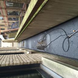 Techmax - Czyszczenie ciśnieniowe zewnętrznej ściany budynku z szarego, chropowatego materiału; widoczny fragment lancy, kabel i mokre ślady po czyszczeniu, ceglana elewacja budynku w tle.