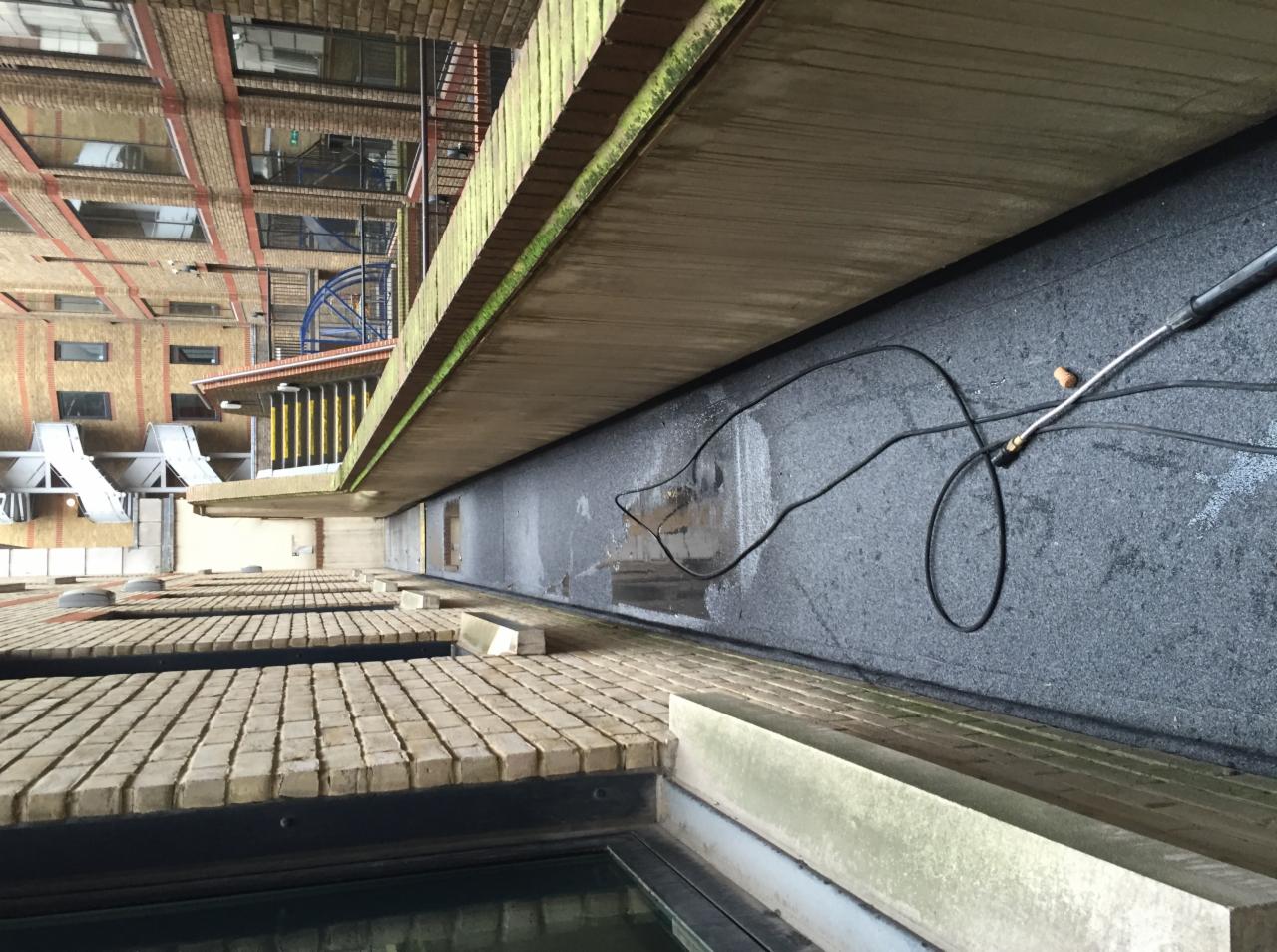 Czyszczenie ciśnieniowe zewnętrznej ściany budynku z szarego, chropowatego materiału; widoczny fragment lancy, kabel i mokre ślady po czyszczeniu, ceglana elewacja budynku w tle.