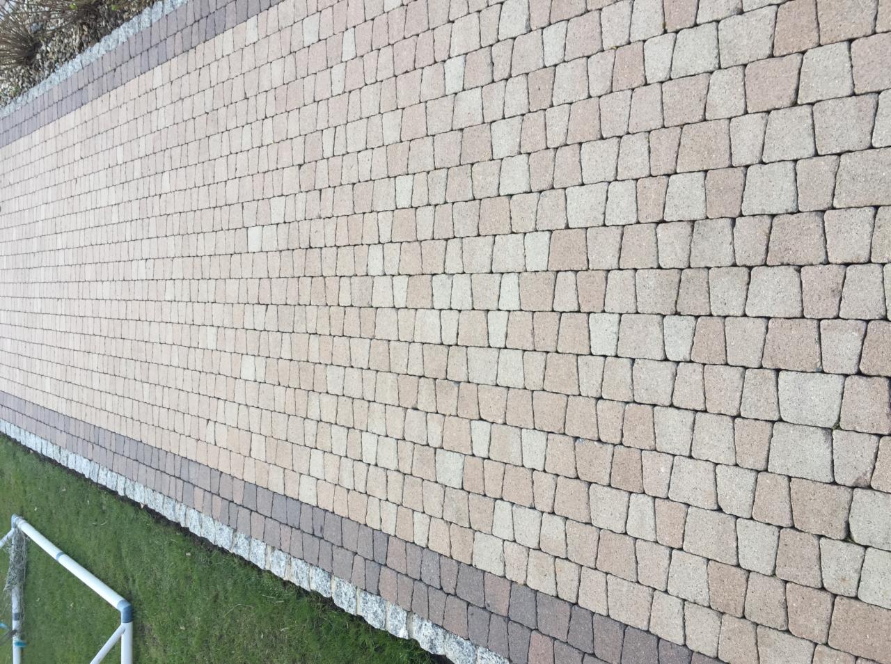 Ułożona kostka brukowa w odcieniach beżu i brązu, obrzeże z ciemniejszej kostki i kamieni, widoczny fragment trawnika i rury PCV.