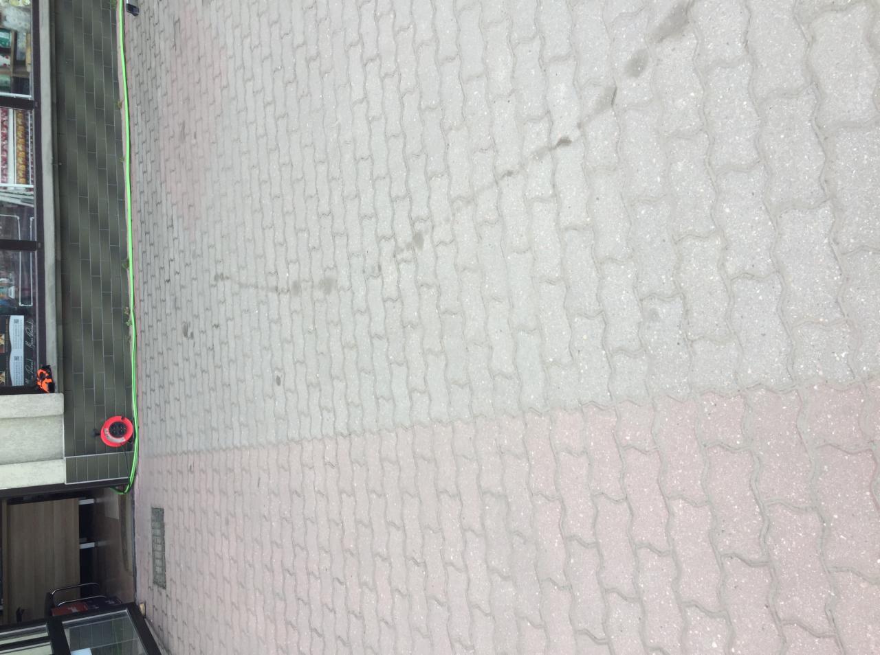 Pusta powierzchnia chodnika z kostki brukowej w odcieniach szarości i różu, widziana z góry, z fragmentem elewacji budynku po lewej stronie i śladami zabrudzeń.