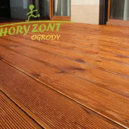 Taras z modrzewia syberyjskiego, deski ryflowane, widoczne okno i fragment elewacji budynku w tle, logo firmy Horyzont Ogrody w lewym górnym rogu.