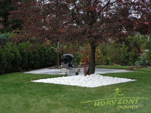 Ogród z drzewem o purpurowych liściach, otoczonym kwadratem z białych kamieni, trawnik, grill na tarasie w tle, logo firmy ogrodniczej w rogu.