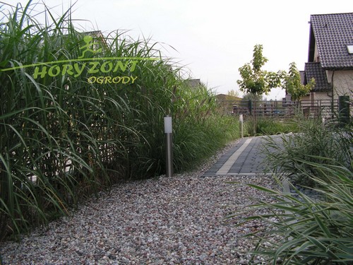 Ścieżka z jasnych kamieni w ogrodzie, obramowana wysokimi trawami ozdobnymi i niskimi lampami oświetleniowymi, widoczne budynki w tle.