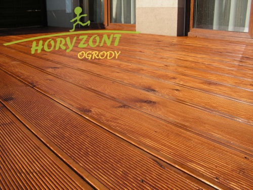 Taras z modrzewia syberyjskiego, deski ryflowane, widoczne okno i fragment elewacji budynku w tle, logo firmy Horyzont Ogrody w lewym górnym rogu.
