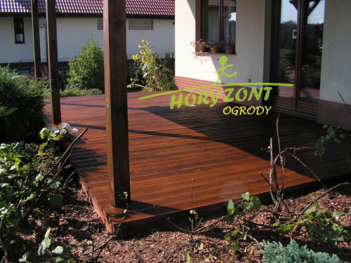 Gotowy taras z modrzewia syberyjskiego przylegający do domu, z drewnianą konstrukcją nośną i rabatami z roślinami ozdobnymi wokół