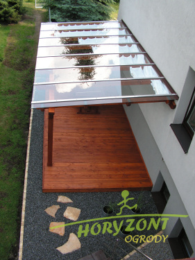 Zadaszony taras szklany przylegający do budynku, wykończony drewnianą podłogą i otoczony czarnym żwirem, z widocznym logo firmy 'Horyzont Ogrody' w prawym dolnym rogu.