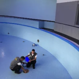 Dwóch mężczyzn w szarych kamizelkach instaluje oświetlenie w pustym basenie z jasnoniebieskimi płytkami i ciemnoniebieskimi akcentami, widok z góry.