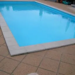 Prostokątny basen z błękitną wodą otoczony jasnoszarą obwódką z kostki, przylegający do brązowej kostki brukowej na zewnątrz, z zwiniętym niebieskim wężem ogrodowym w lewym dolnym rogu.