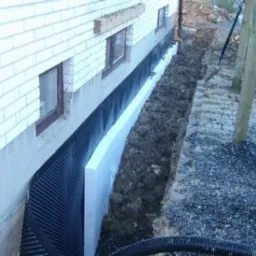 Fundament budynku z białej cegły po izolacji pionowej: czarna folia kubełkowa i biały styropian w wykopie.