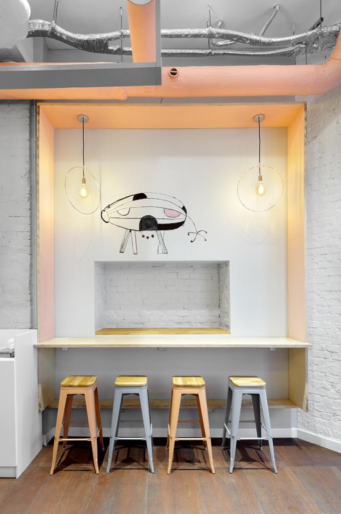 Kącik barowy w biurze z czterema stołkami, rysunkiem na ścianie i wiszącymi lampami z żarówkami Edisona; widoczne rury wentylacyjne pod sufitem.