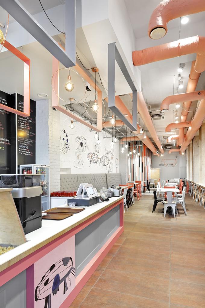 Wnętrze restauracji z industrialnym designem: widoczne rury wentylacyjne pomalowane na różowo pod sufitem, czarno-biała grafika na ścianie z cegły, szare i różowe elementy konstrukcyjne pod sufitem...